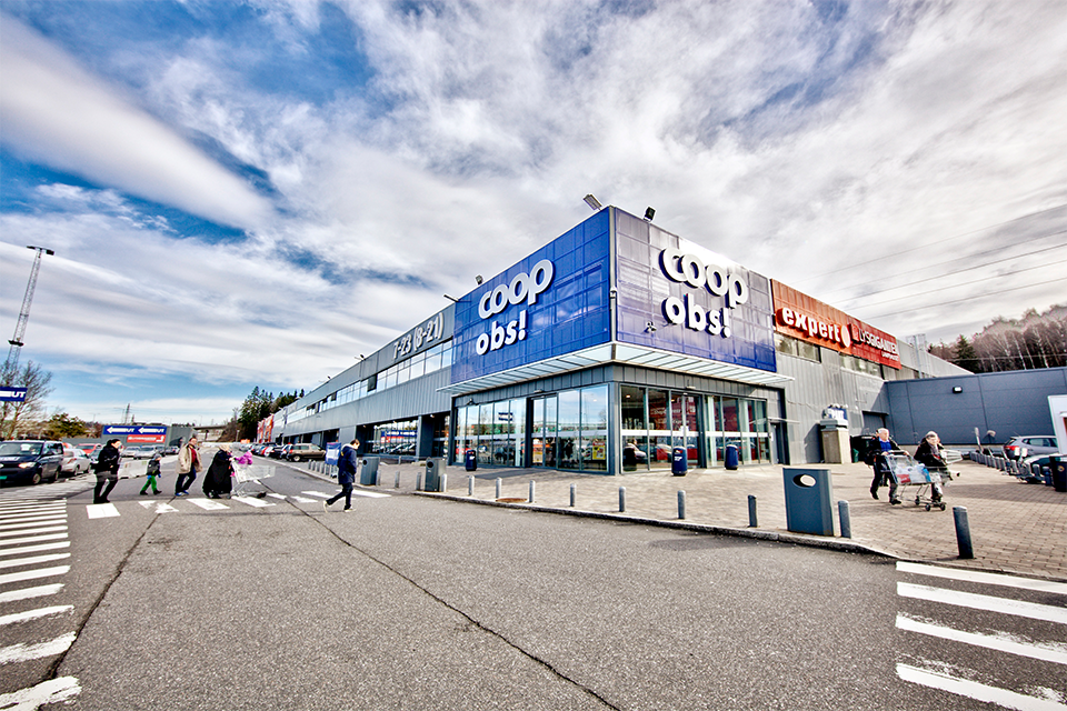 The outside of a Coops Norge grocery store in Norway, a tenant of W. P. Carey. Several shoppers are walking into the building, which has lots of glass windows and a large blue and white sign saying "Coop obs!".